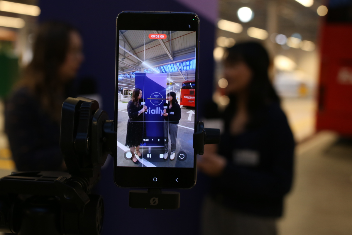 A smartphone mounted on a tripod displays a video recording in progress. The phone screen shows two people standing in front of a navy blue 'vially' banner in a large, brightly lit transit station with a red bus in the blurred background.
